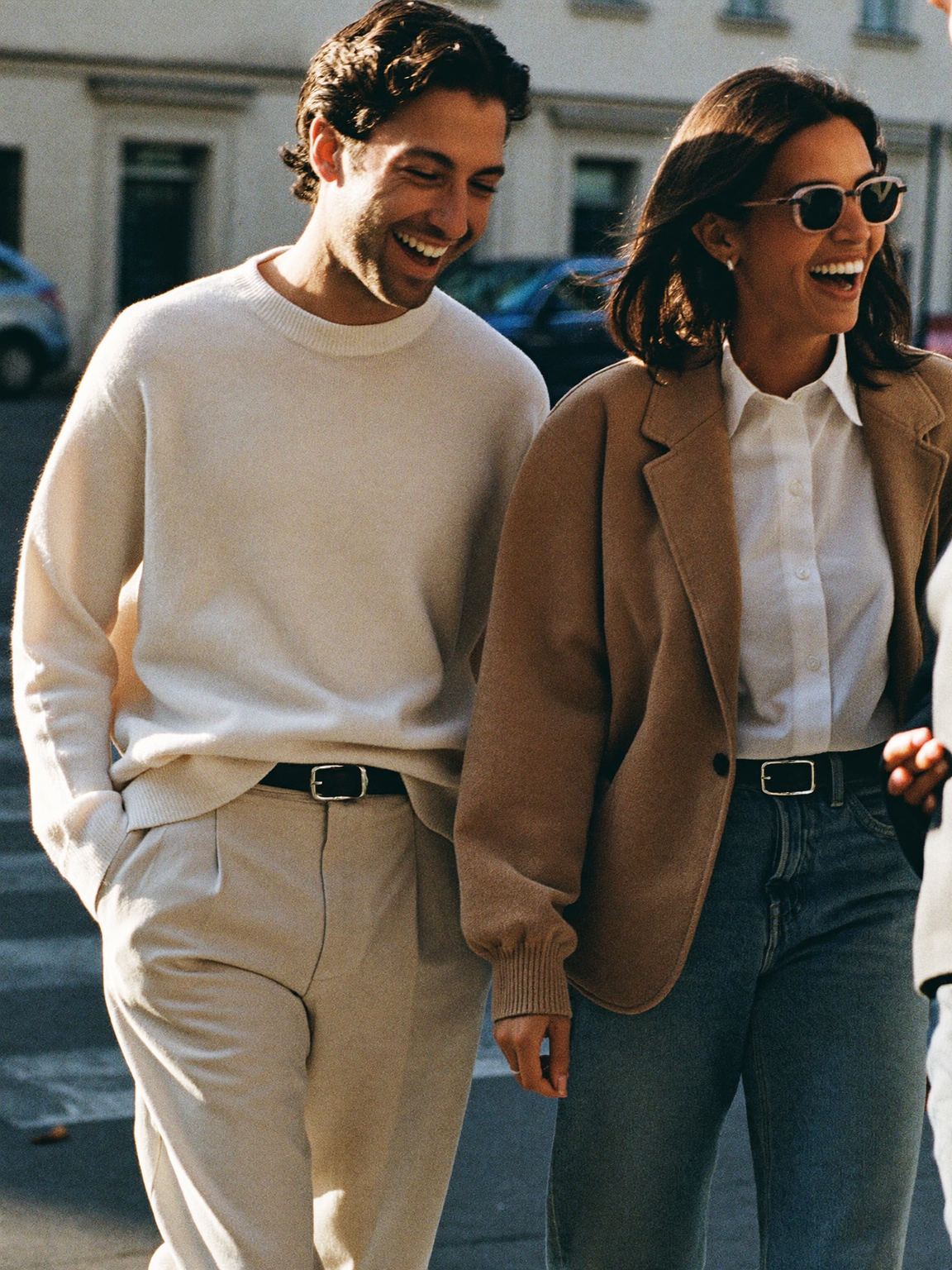 Two friends laughing together on a sunlit street in relaxed, premium-casual outfits